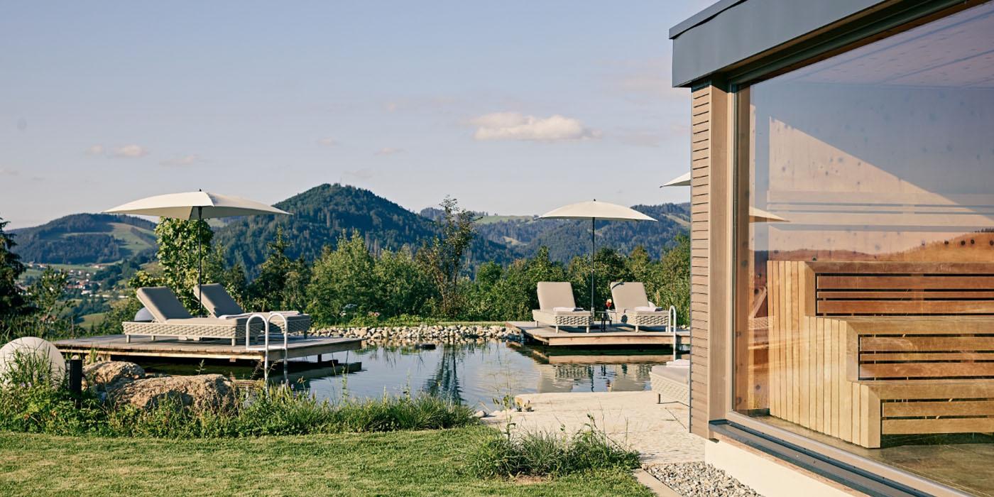 Natur-Schwimmteich mit Holzdecks und Liegen vor der Panoramasauna des Hotel Birkenhof Oberstaufen, mit Blick auf die Allgäuer Berge.