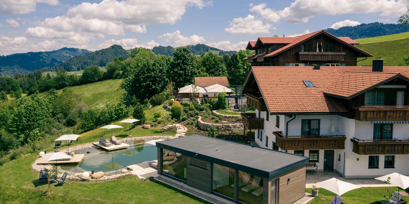 Außenansicht des Hotel Birkenhof Oberstaufen mit Natur-Schwimmteich, Liegebereich und Blick auf die umliegende Allgäuer Hügellandschaft.